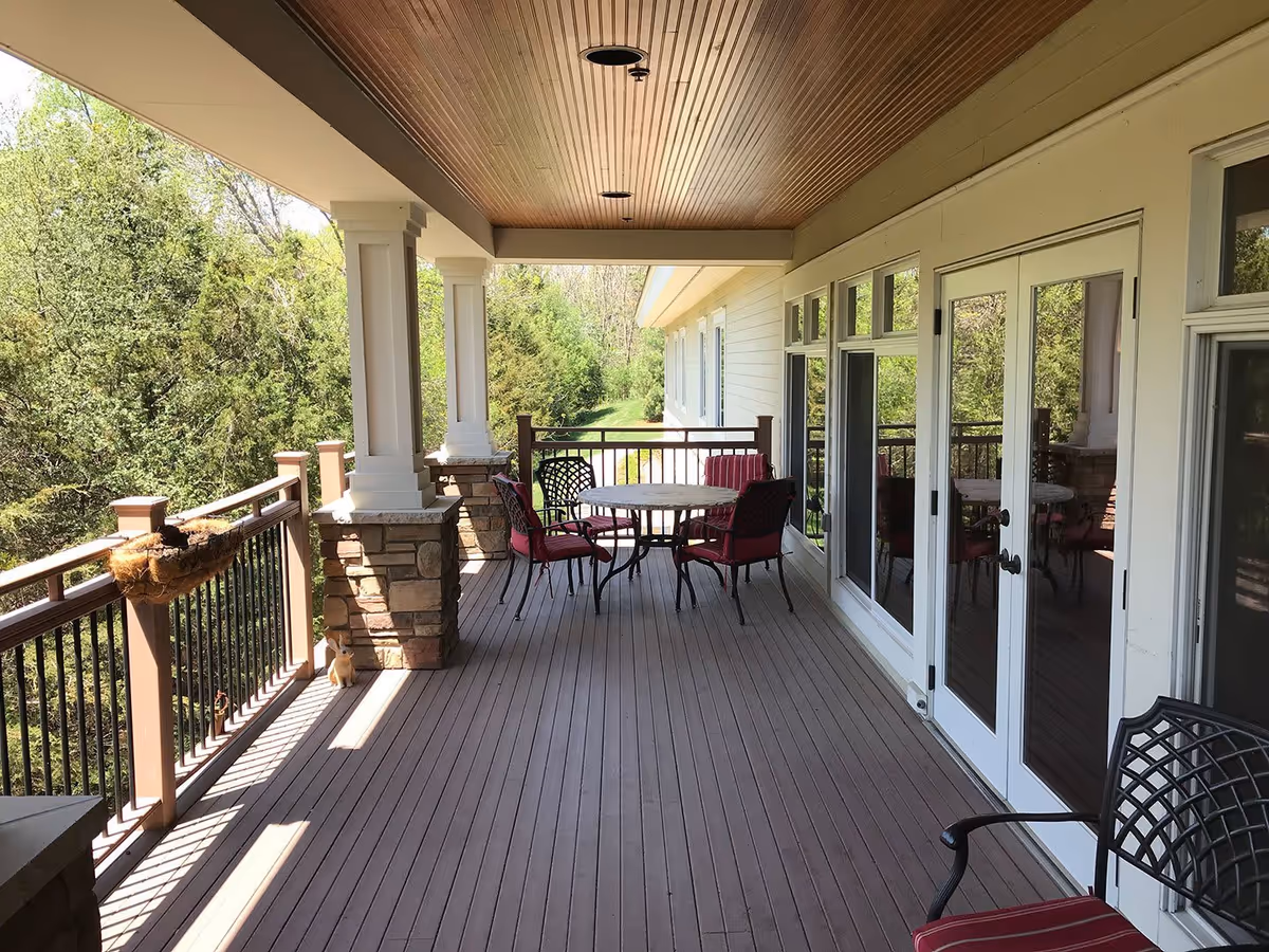 Covered outdoor deck with a round table and red-cushioned chairs overlooking trees.