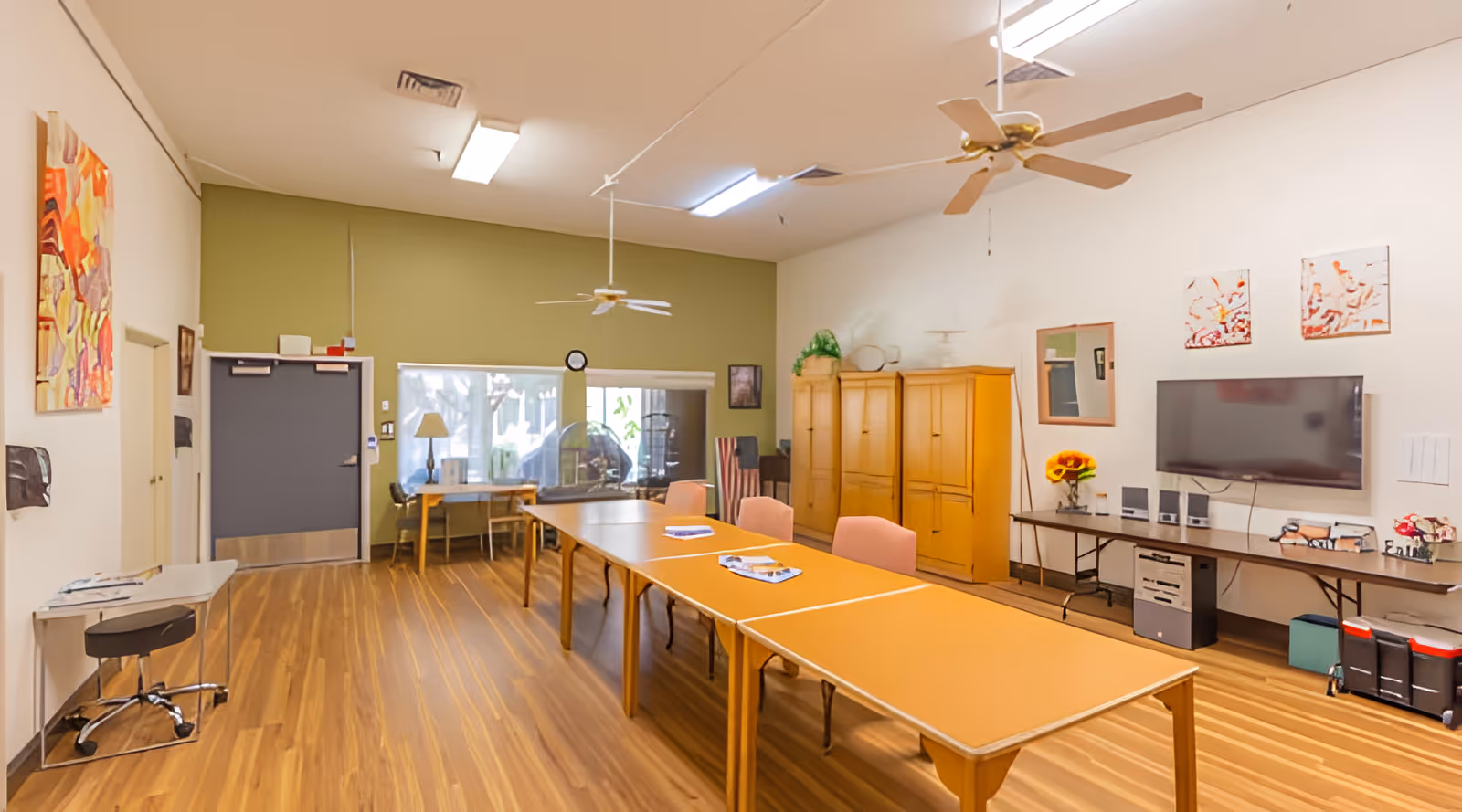Bright activity room with long tables and chairs, a wall-mounted TV, wooden cabinets, and ceiling fans.