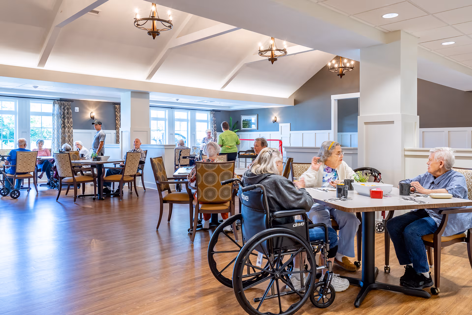 Elderly residents sit and converse around tables in a bright, spacious communal dining room.