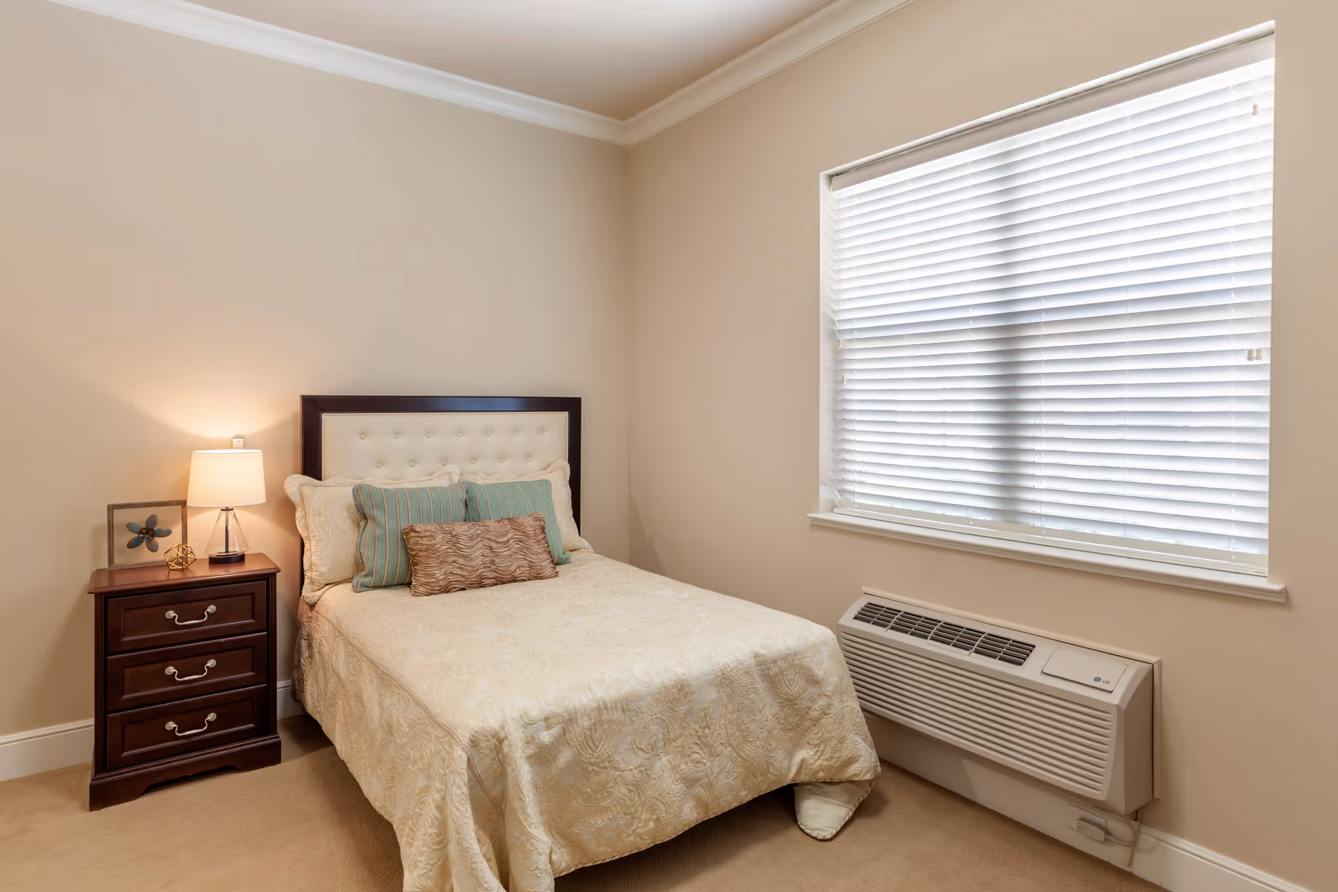 A small bedroom with a neatly made bed featuring a cream-colored patterned bedspread and multiple pillows in beige, green, and brown tones. Next to the bed is a dark wooden nightstand with three drawers, a lamp, and decorative items. A window with white blinds is on the right wall above a wall-mounted air conditioning unit.