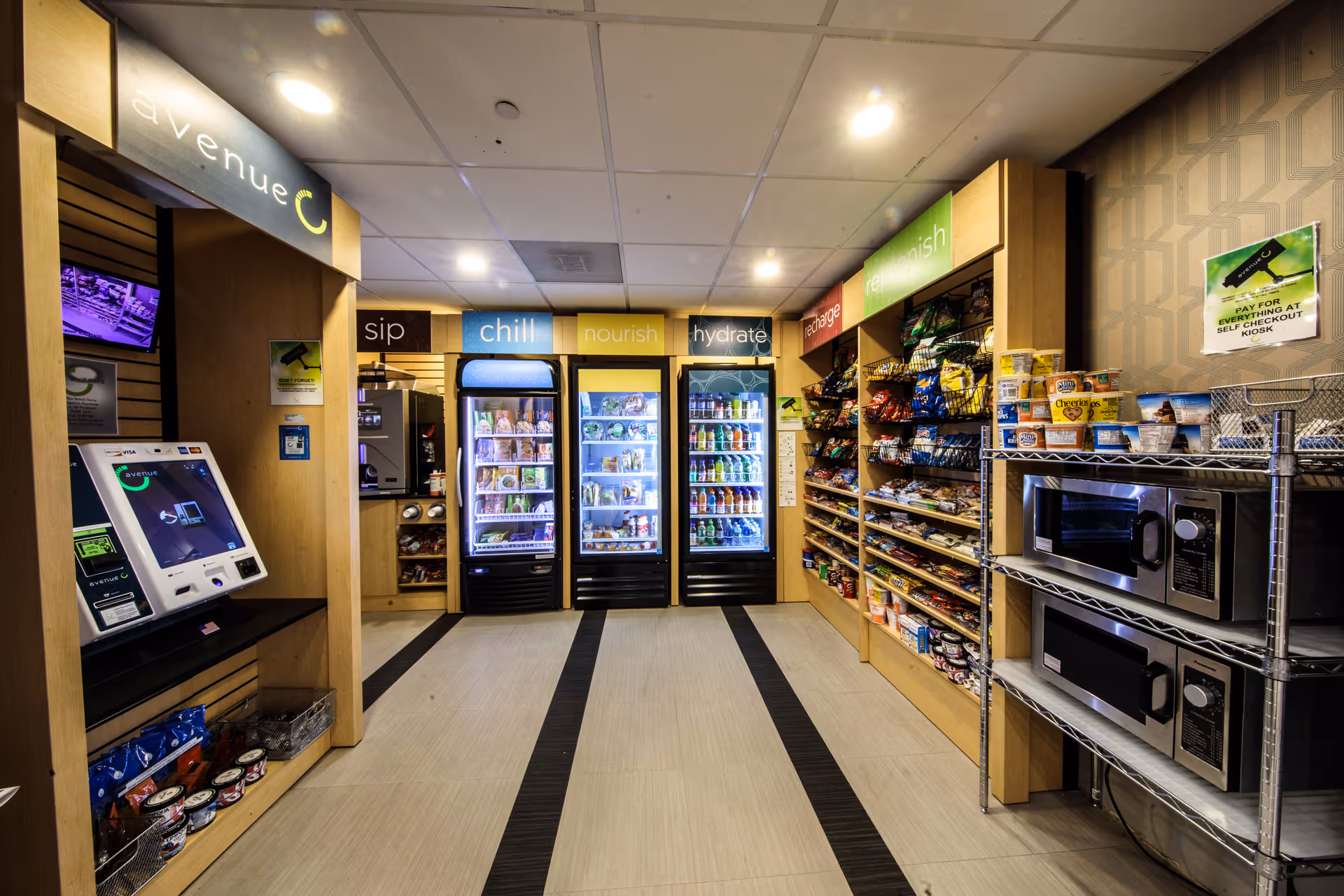 Interior view of a small convenience store area inside a facility, featuring refrigerated display cases labeled chill, nourish, and hydrate, shelves stocked with snacks, two microwaves on a metal rack, and a self-checkout kiosk on the left side under a sign that reads avenue C.