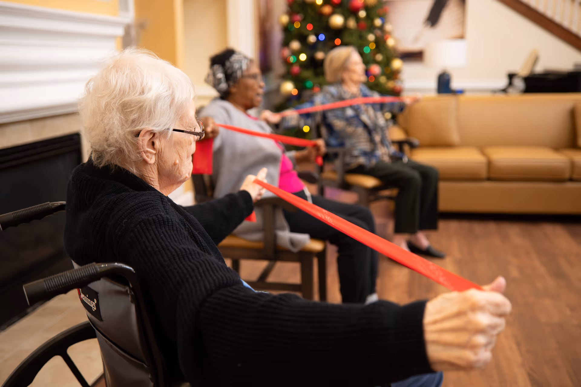Three elderly women seated indoors in a living room area, exercising with red resistance bands. A decorated Christmas tree is visible in the background along with a tan couch and wooden flooring.