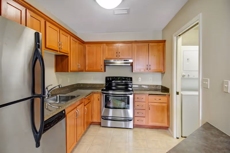 Modern kitchen with wooden cabinets, granite countertops, stainless steel refrigerator, stove, and dishwasher. There is a double sink and a small laundry area with a stacked washer and dryer visible through an open door.