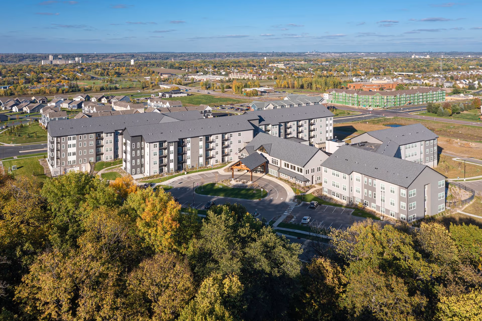 Aerial view of a large senior living community building complex surrounded by trees with autumn foliage. The multi-story buildings have gray roofs and light-colored walls. There is a circular driveway with a covered entrance in the center. The surrounding area includes roads, parking lots, and other buildings in the distance under a partly cloudy blue sky.