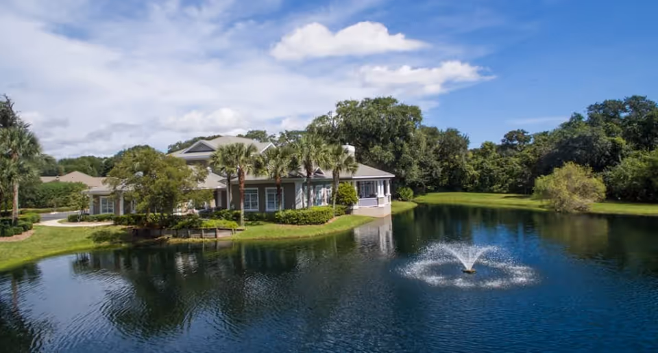 A serene outdoor scene featuring a building surrounded by lush greenery and palm trees, situated next to a pond with a water fountain spraying water into the air under a partly cloudy sky.