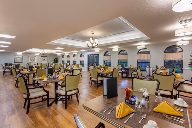 Spacious senior living dining room with multiple set tables, yellow napkins, green-upholstered chairs, chandeliers, and arched windows.