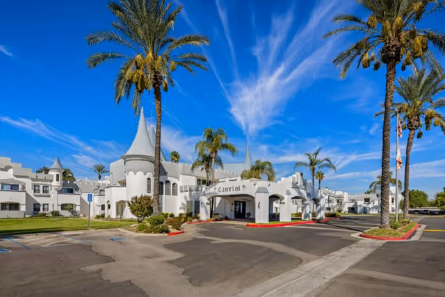 Exterior view of Camelot - A Provincial Senior Living Community featuring a white building with castle-like architectural elements including turrets and battlements, surrounded by palm trees under a bright blue sky with wispy clouds. The entrance has a covered driveway with the name 'Camelot' displayed above it.