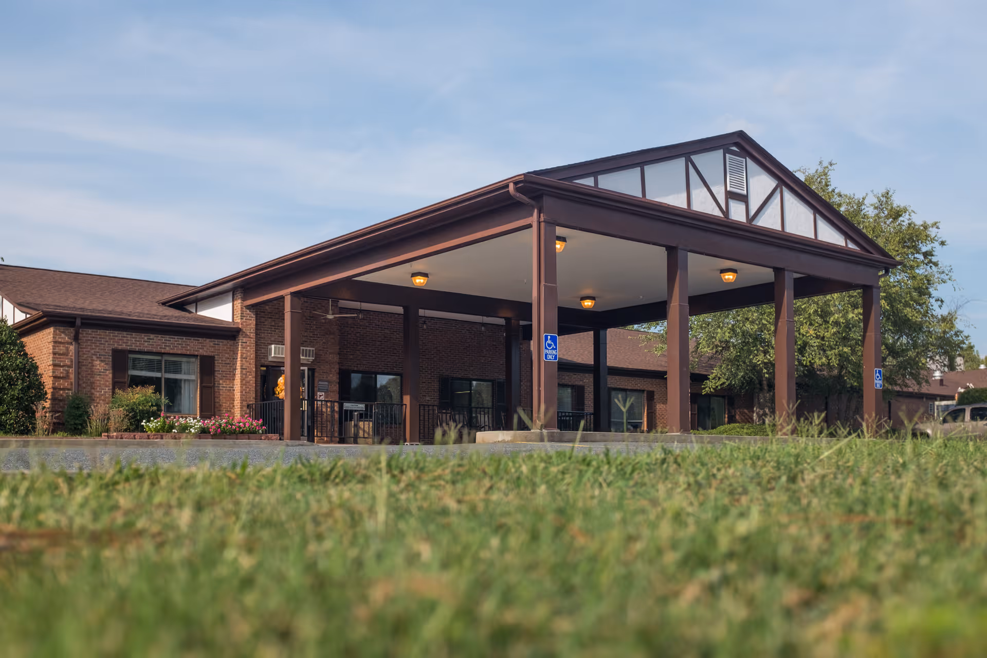 Exterior view of White Oak Manor Lancaster showing the front entrance with a covered drop-off area supported by brown pillars, brick walls, windows, and a well-maintained lawn in the foreground under a blue sky.