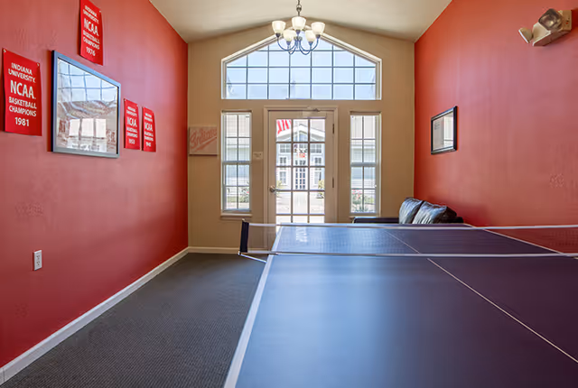 Interior room with red walls featuring Indiana University NCAA basketball championship banners, a ping pong table in the foreground, a black leather couch against the right wall, and a glass door with windows letting in natural light.