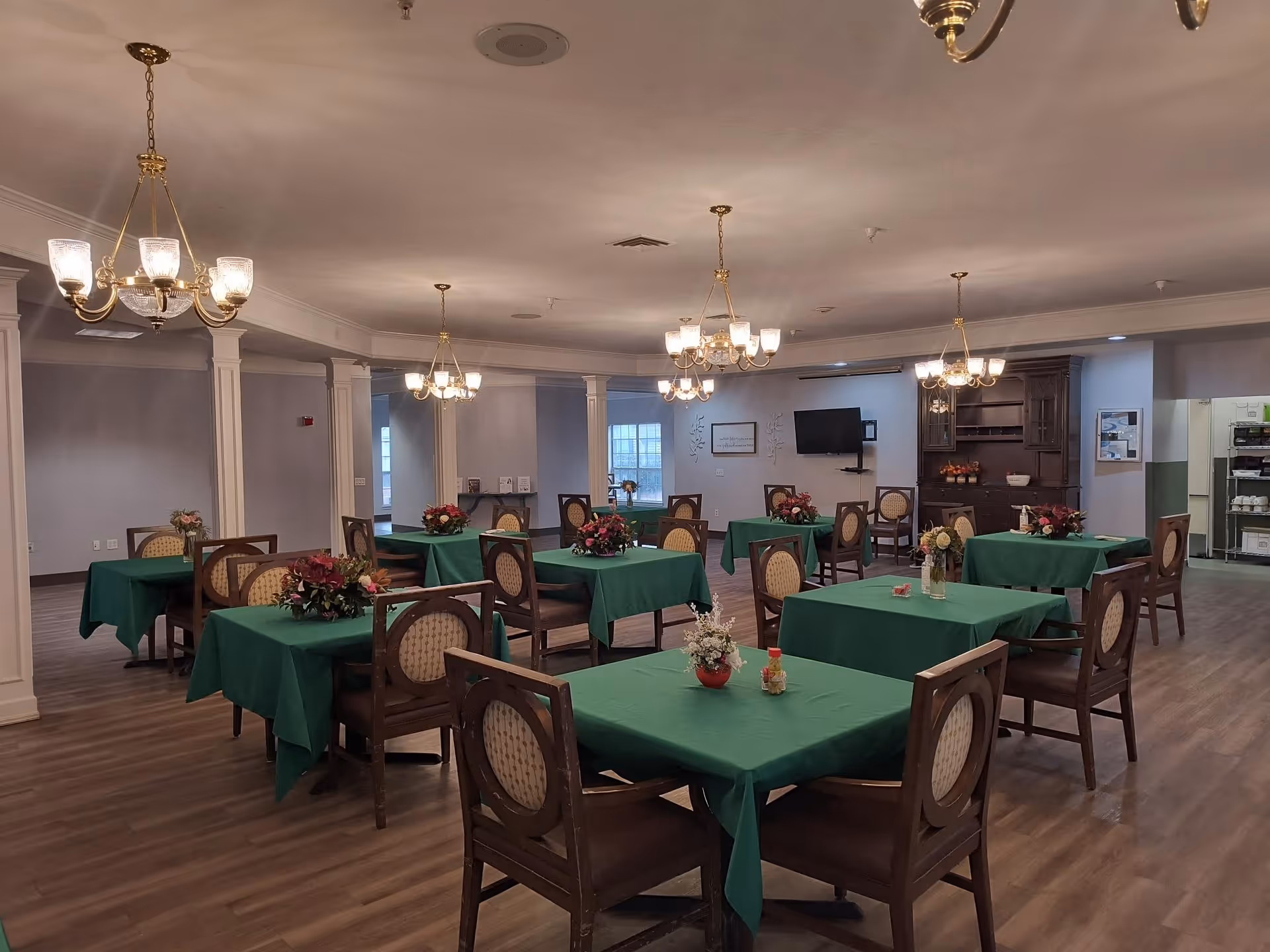 A dining room in a senior living facility with multiple tables covered in green tablecloths, each adorned with floral centerpieces. The room features wooden chairs with cushioned backs, chandeliers hanging from the ceiling, and a wooden cabinet against the far wall. The floor is wood, and there is a television mounted on the wall.