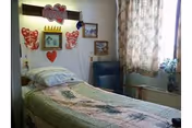 A small bedroom with a single bed covered with a patterned quilt. Above the bed, there are decorative wall hangings including hearts and a sign that says 'Love'. Next to the bed is a blue chair and a window with floral curtains letting in natural light. A small plant is placed on the windowsill.