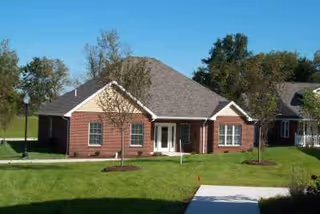 Single-story brick senior living building with a pitched roof, white-trimmed entrance, and landscaped lawn under a clear blue sky.