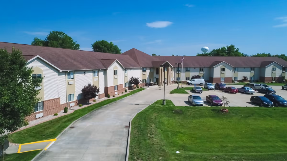 Two-story senior living building with a front driveway, parking lot, flagpole, and green lawn under a blue sky.