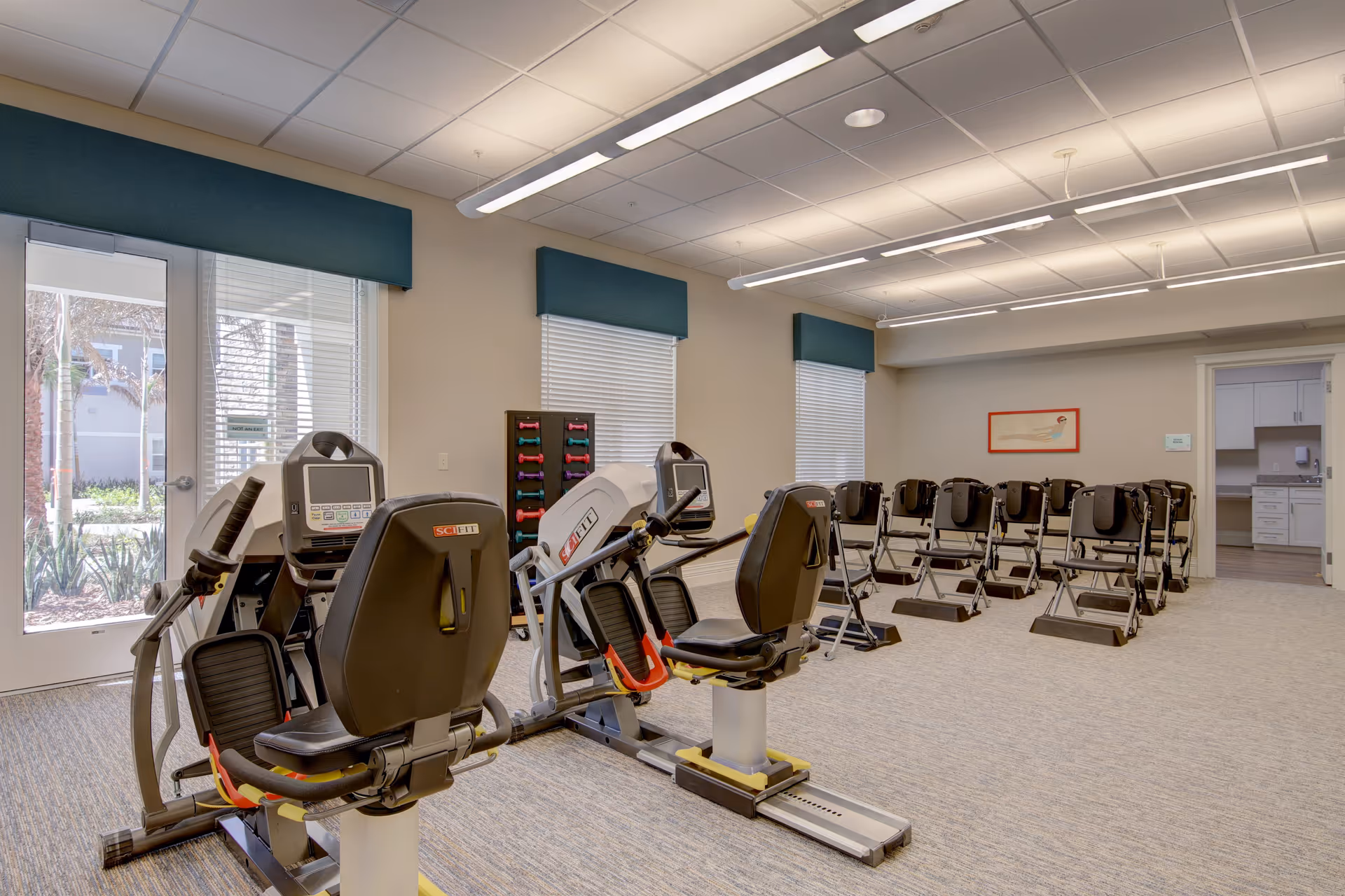 A well-lit exercise room with multiple recumbent exercise bikes and a rack of colorful dumbbells against the wall. The room has large windows with blinds and teal valances, and a door leading outside. There are also several black chairs arranged in rows facing the exercise equipment. The ceiling has fluorescent lighting and the floor is carpeted.
