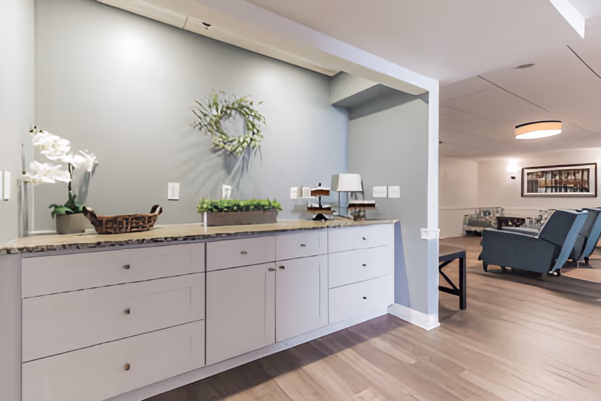 Interior view of a senior living facility showing a countertop with white cabinets and decorative items including a plant, a basket, and a lamp. In the background, there is a seating area with armchairs, a coffee table, and a framed picture on the wall. The room has wooden flooring and soft lighting.