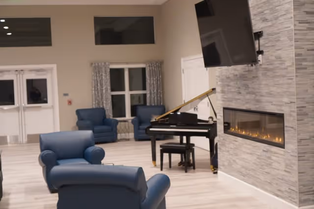 A spacious living room area with light wood flooring, featuring several blue armchairs, a black grand piano with a matching bench, a modern electric fireplace set in a gray tiled wall, and a large flat-screen TV mounted above the fireplace. There are windows with patterned curtains and double doors in the background.
