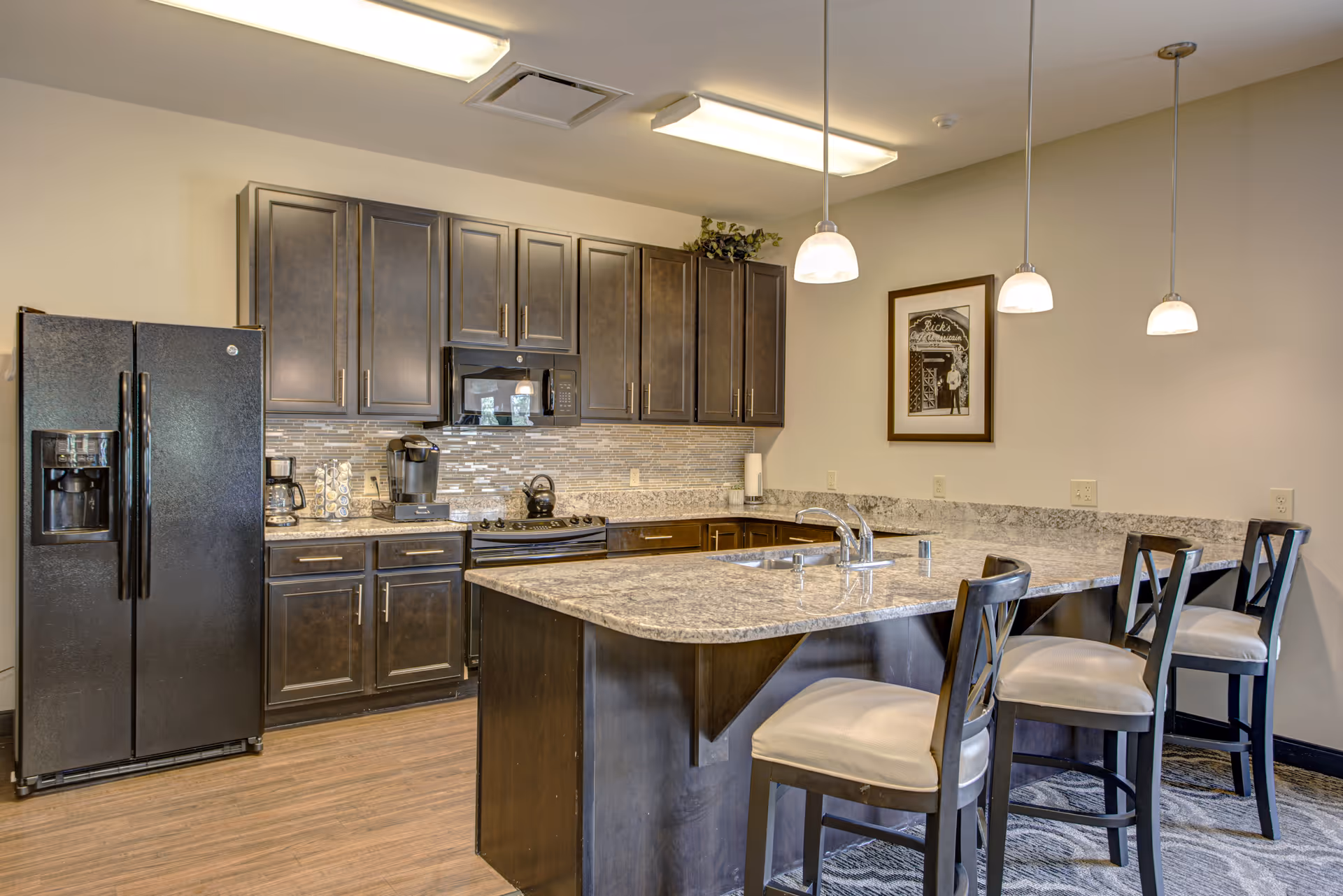Modern kitchen area with dark wood cabinets, a black refrigerator, a stove with a microwave above it, a coffee maker, and a large granite countertop island with a sink and four cushioned chairs. Three pendant lights hang above the island, and a framed picture is on the wall.