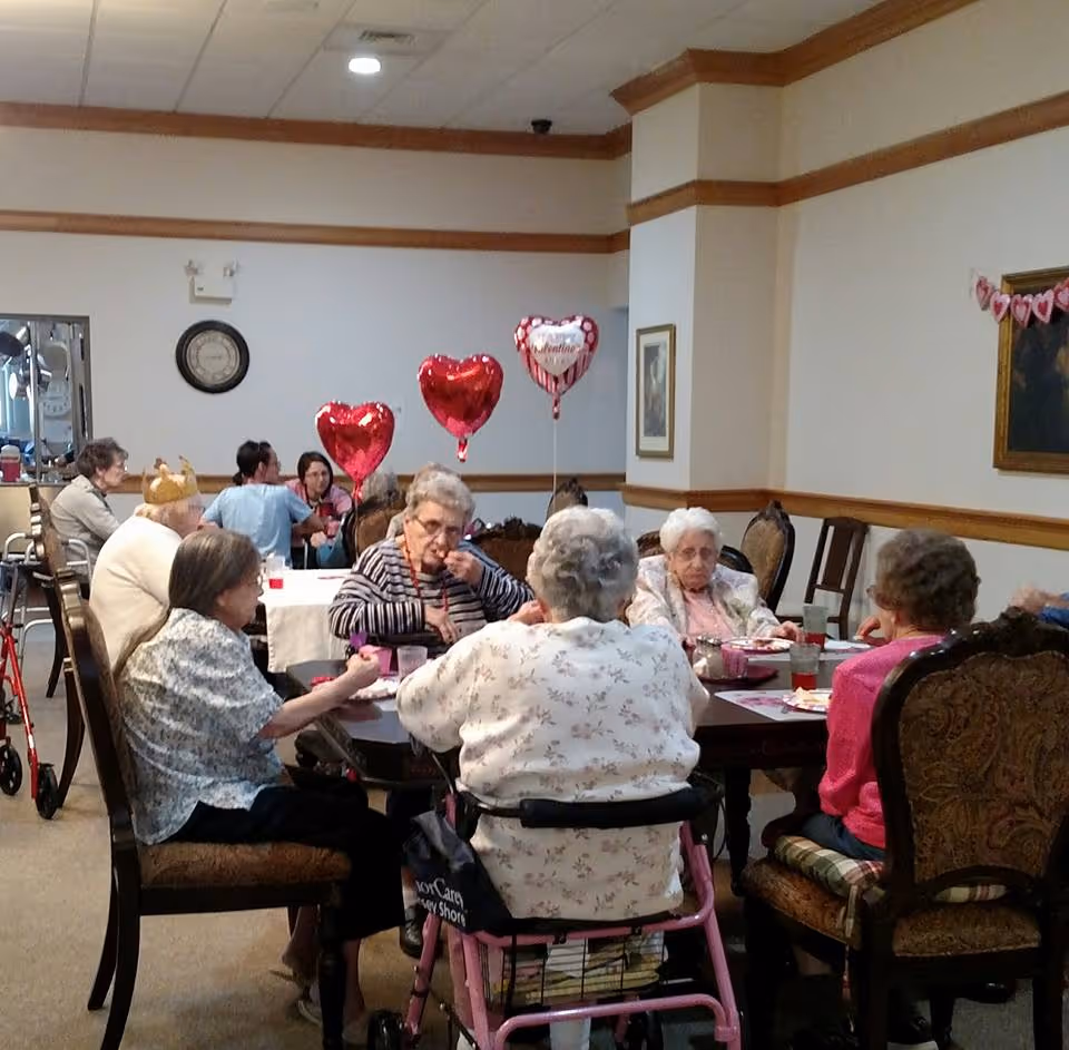 A group of elderly residents seated around tables in a decorated dining/activity room with heart-shaped balloons.