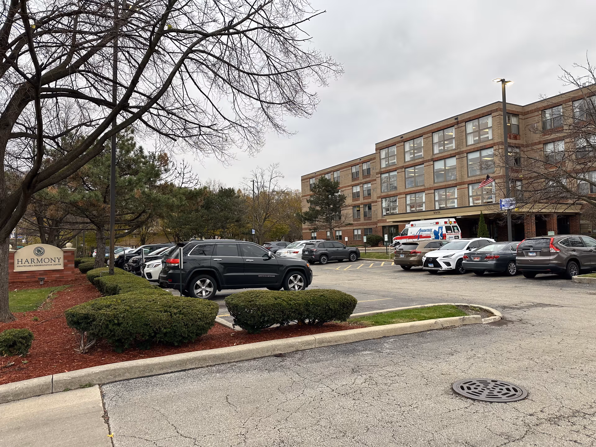 Exterior view of Harmony Healthcare & Rehabilitation Center showing a multi-story brick building with many windows, a parking lot filled with cars, an ambulance parked near the entrance, and landscaped bushes and trees under a cloudy sky.