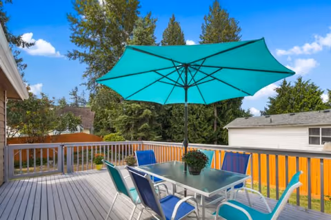 Outdoor wooden deck with a glass-top table surrounded by six colorful chairs and a large teal patio umbrella. The deck is enclosed by a railing, and there are trees and a wooden fence in the background under a blue sky with some clouds.