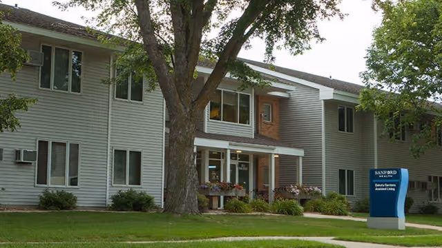 Two-story light-gray residential building with a large tree and a Sanford Health sign near the front entrance.