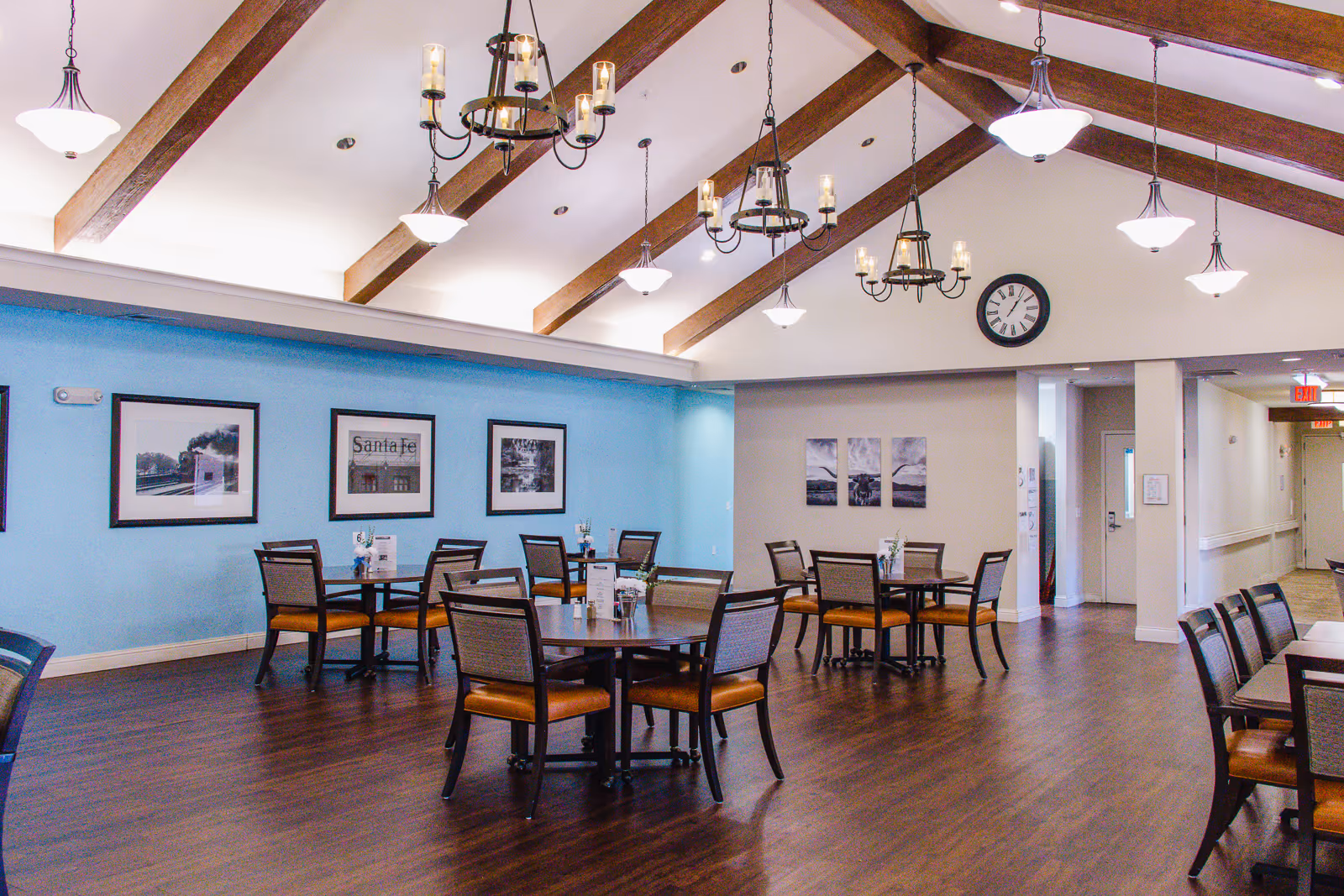 Bright assisted living dining room with round tables and chairs under a vaulted beamed ceiling and chandeliers.