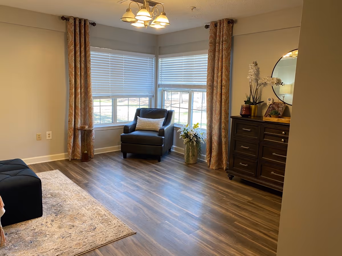 A cozy living room corner with a dark armchair featuring a white cushion, positioned near two large windows with white blinds and patterned curtains. A wooden dresser with drawers is on the right side, decorated with a round mirror, a lamp, and flower arrangements. The floor is wood with a light-colored area rug partially visible.