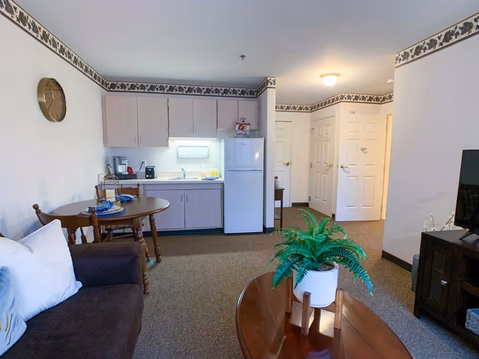 Interior view of a small assisted living apartment featuring a kitchenette with light-colored cabinets, a white refrigerator, and a sink. A round wooden dining table with two chairs is set with blue napkins and plates. A brown sofa with white pillows is partially visible on the left. A wooden coffee table with a potted green plant is in the foreground. The room has beige carpet, white walls with a floral border near the ceiling, and a wall clock. There is a TV on a dark wooden stand on the right side and two white doors in the background.