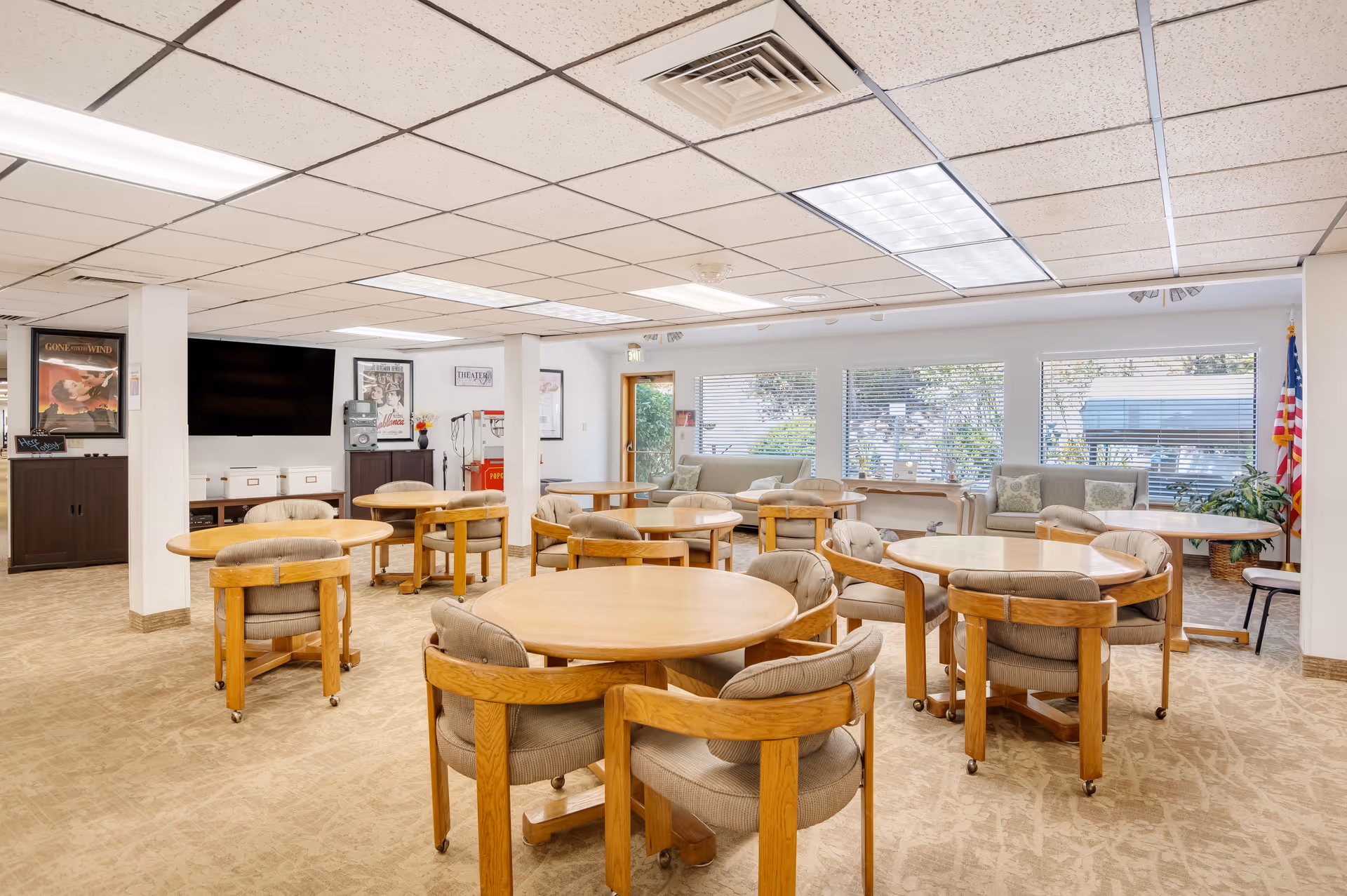 A bright and spacious common area with multiple round wooden tables and cushioned chairs on wheels. The room has large windows with blinds letting in natural light, a flat-screen TV mounted on the wall, movie posters, a popcorn machine, and comfortable seating along the windows. The carpet is beige with a subtle pattern, and the ceiling has fluorescent lighting panels.
