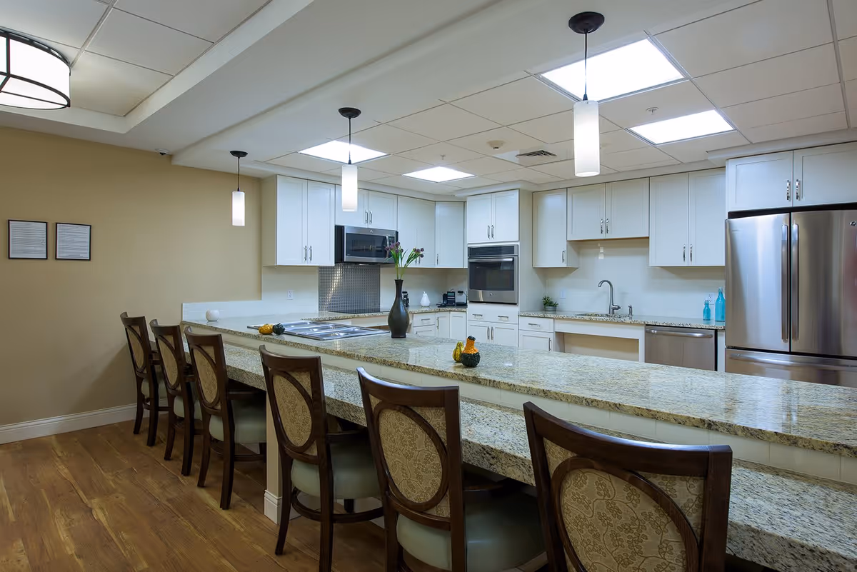 Modern kitchen with a long granite countertop island featuring six wooden chairs with cushioned seats. The kitchen has white cabinets, stainless steel appliances including a refrigerator, oven, microwave, and dishwasher. Pendant lights hang from the ceiling, and the floor is wooden.