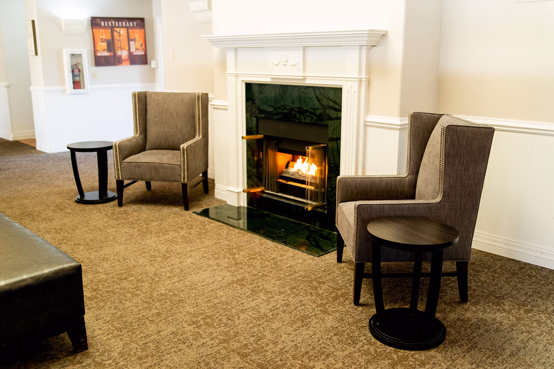 A cozy seating area in a senior living facility with two gray upholstered armchairs, each accompanied by a small round black side table, positioned on a brown carpet in front of a white fireplace with a fire burning inside. The walls are light-colored with white wainscoting, and a framed picture hangs on the wall in the background.