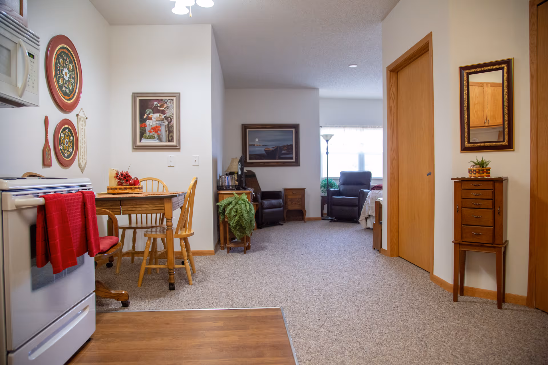 Interior view of a senior living apartment showing a small kitchen area with a stove and microwave on the left, a wooden dining table with chairs, and a living area with two armchairs and a floor lamp near a window in the background. The walls are decorated with framed pictures and decorative plates. There is a small wooden cabinet with a mirror above it on the right side.