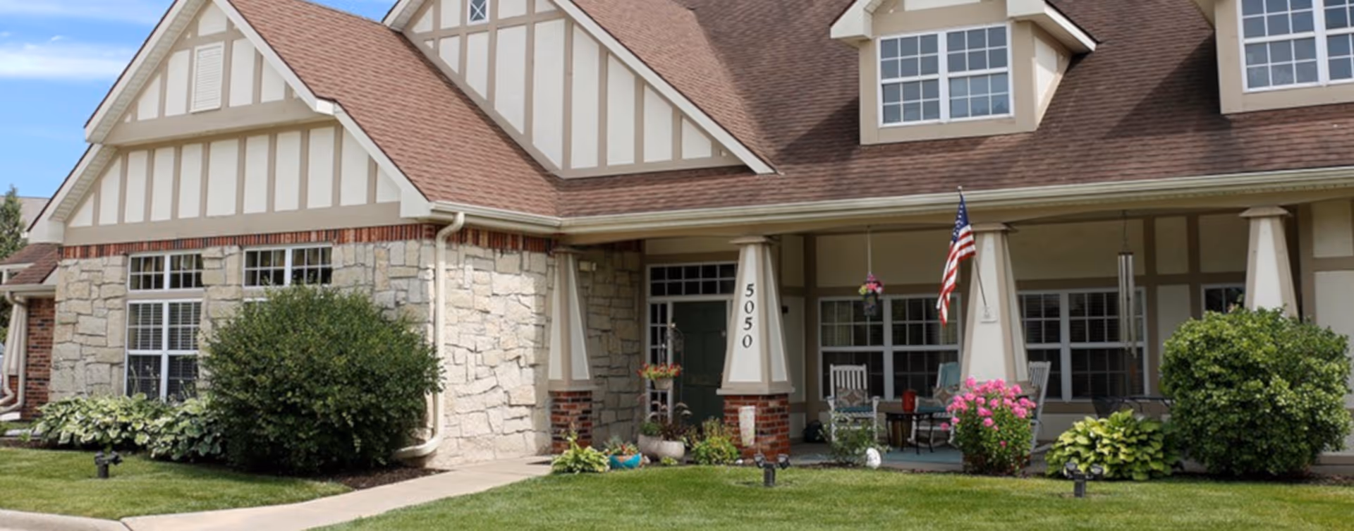 Front exterior view of a senior living facility building with stone and beige siding, a covered porch with rocking chairs, potted plants, an American flag, and well-maintained lawn and shrubs.