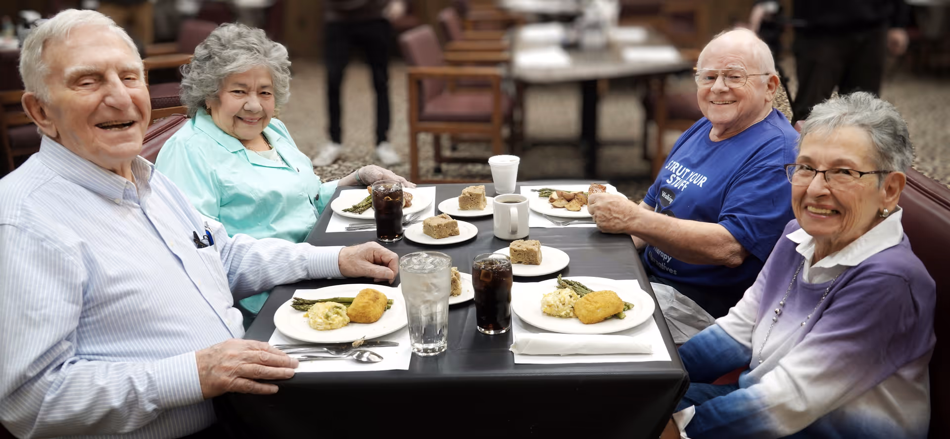 Four elderly people sitting around a dining table in a senior living facility, smiling and enjoying a meal together. The table has plates with food, glasses of water and soda, and coffee cups. The background shows more tables and chairs in a communal dining area.