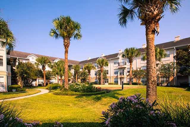 A sunny courtyard with green grass, palm trees, and flowering bushes surrounded by a three-story residential building under a clear blue sky.