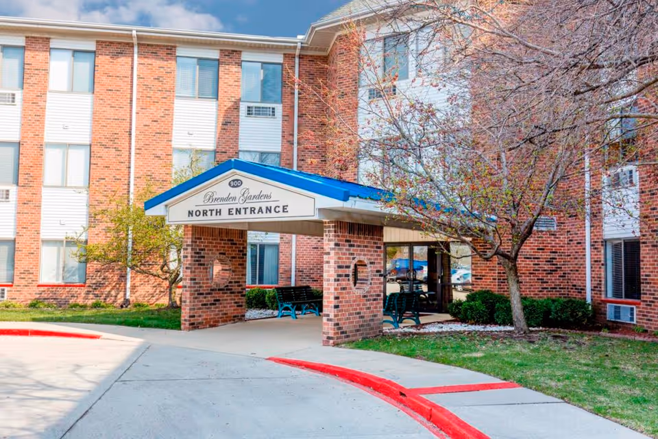Exterior view of Brenden Gardens senior living facility showing the north entrance with a covered walkway supported by brick pillars. There are benches under the entrance canopy, a tree with budding leaves, and a red curb along the driveway.