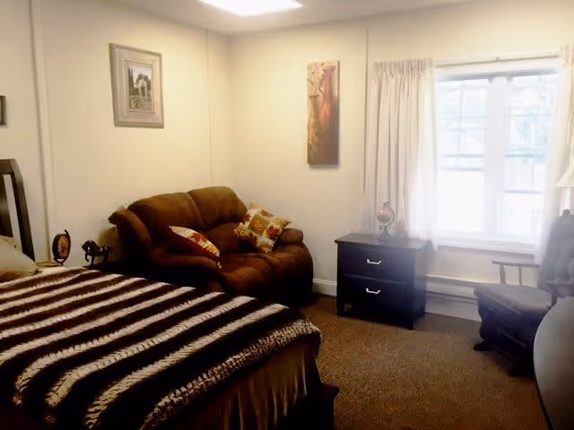 A cozy bedroom with a bed covered in a striped blanket, a brown loveseat with decorative pillows, a nightstand with a small globe, a window with curtains, and a cushioned chair near a round table.