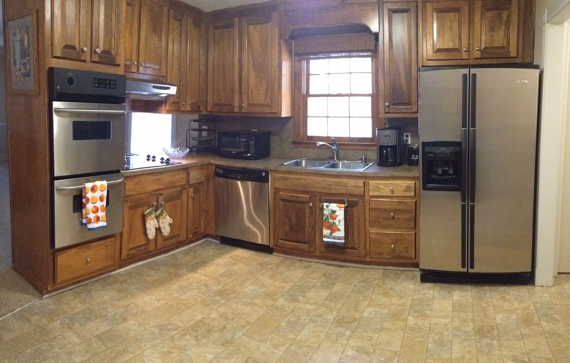 A kitchen with wooden cabinets, a double oven, a dishwasher, a double sink under a window, a coffee maker, a toaster, and a stainless steel refrigerator. The floor has a tile pattern, and there are decorative towels hanging on the oven and cabinet handles.