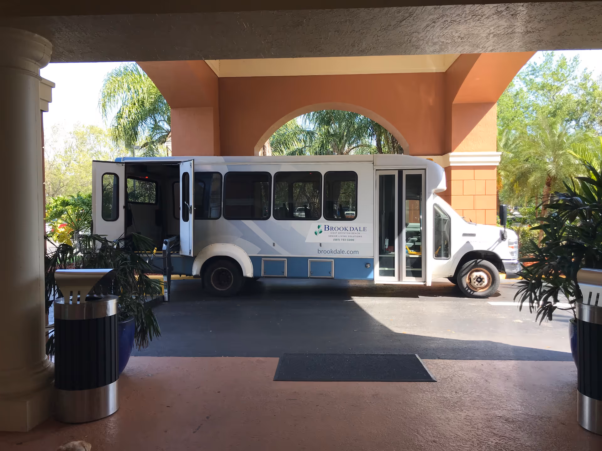 A white Brookdale shuttle bus parked under a covered entrance with its rear door open. The bus has the Brookdale logo and website on its side. There are plants and trash bins on either side of the entrance, and palm trees are visible in the background.