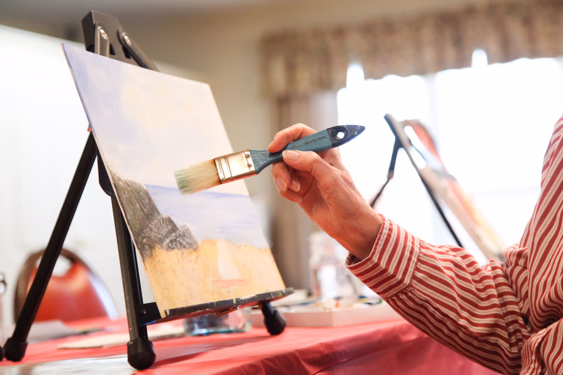 A person wearing a red and white striped shirt is painting a landscape on a canvas set on an easel. The painting depicts a beach scene with sand, water, and rocks. The background shows a softly lit room with red chairs and windows with curtains.