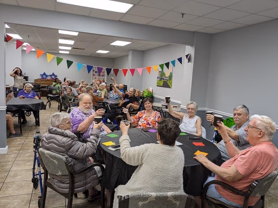A group of elderly people sitting around tables in a decorated room, raising cups in a toast. The room has colorful triangular pennant banners hanging from the ceiling and a birthday sign in the background. The atmosphere appears festive and social.