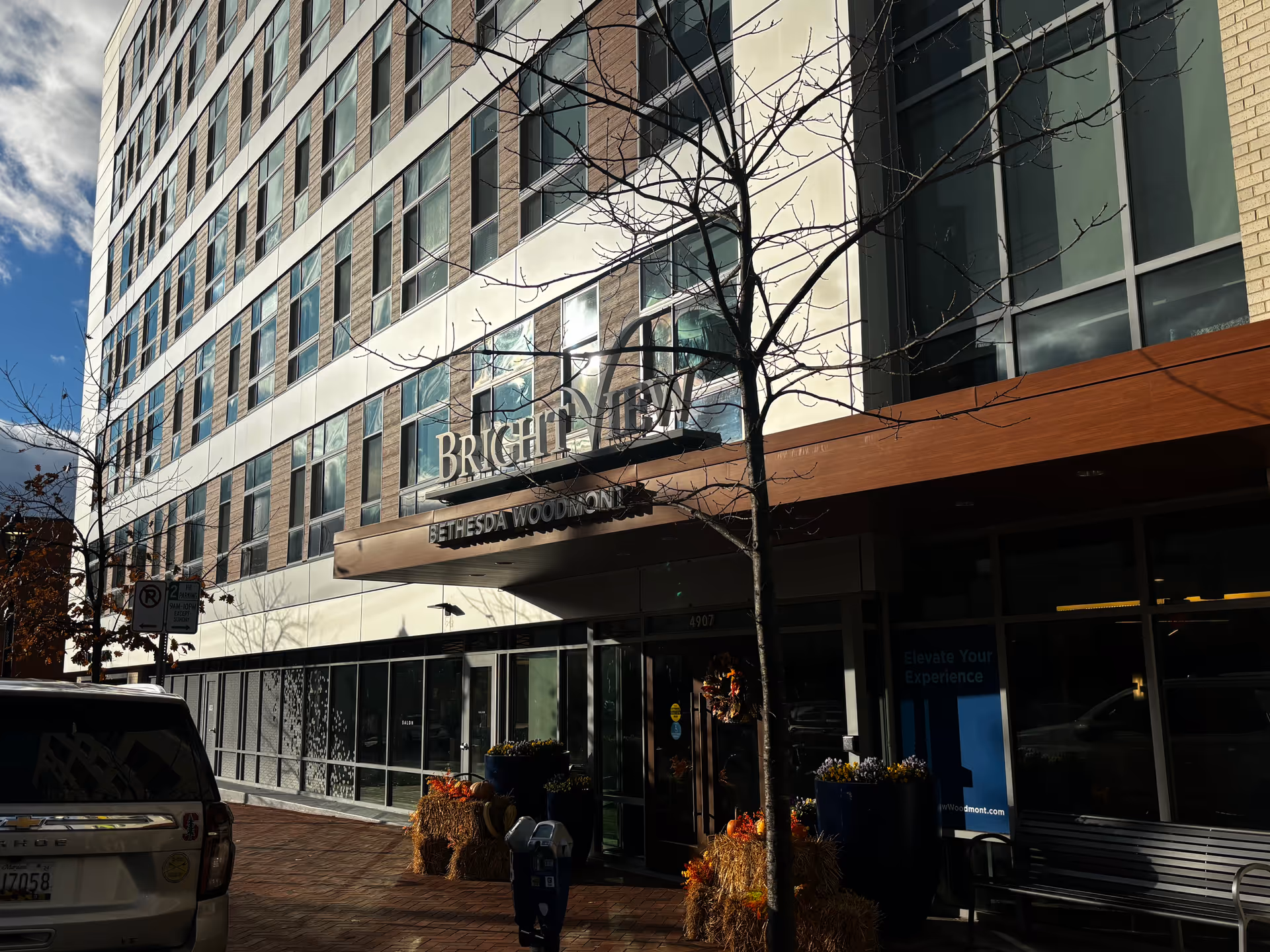 Exterior view of a multi-story building with large windows and a sign that reads 'BRIGHTVIEW BETHESDA WOODMONT' above the entrance. There are some fall-themed decorations including hay bales and pumpkins near the entrance. A parked white Chevrolet vehicle and a parking meter are visible on the brick sidewalk in front of the building. Leafless trees line the sidewalk under a partly cloudy sky.