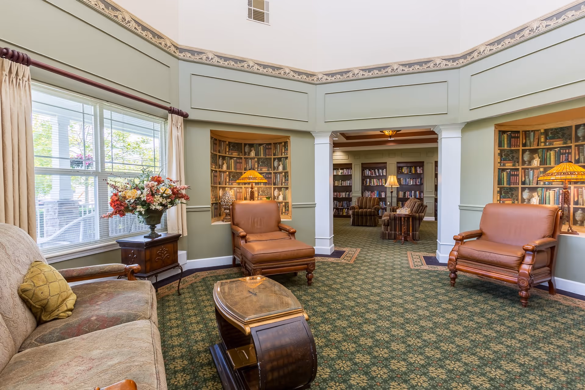 A cozy sitting area in a senior living facility with a beige sofa, two brown leather armchairs, a wooden coffee table, and a side table with a floral arrangement. The room has green patterned carpet, light green walls with decorative molding, and large windows with beige curtains. In the background, there is an adjoining room with bookshelves filled with books and additional seating.