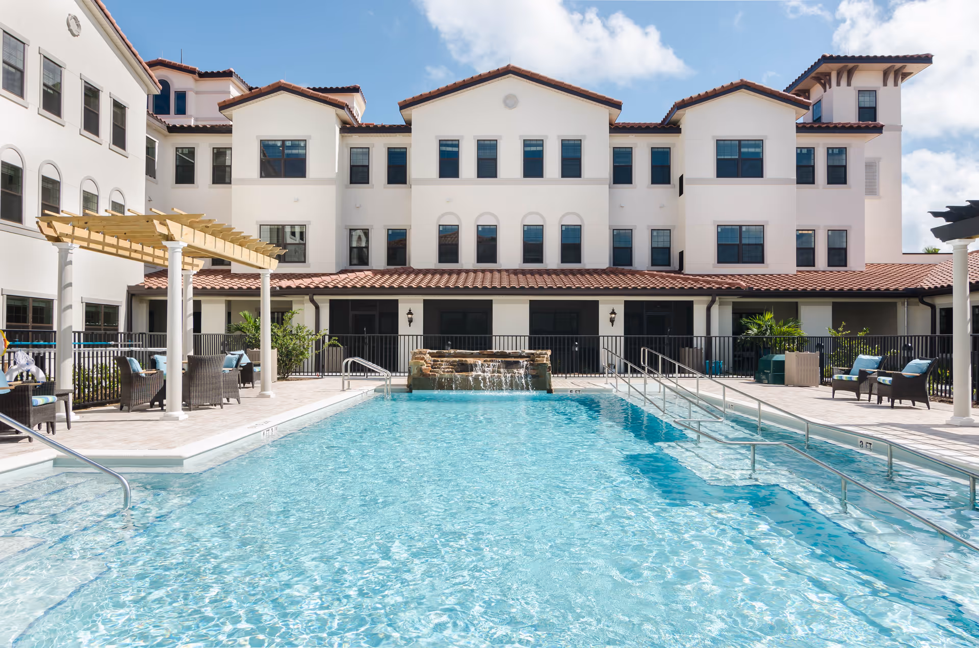A clear blue outdoor swimming pool with lounge seating and a Mediterranean-style multi-story building in the background.