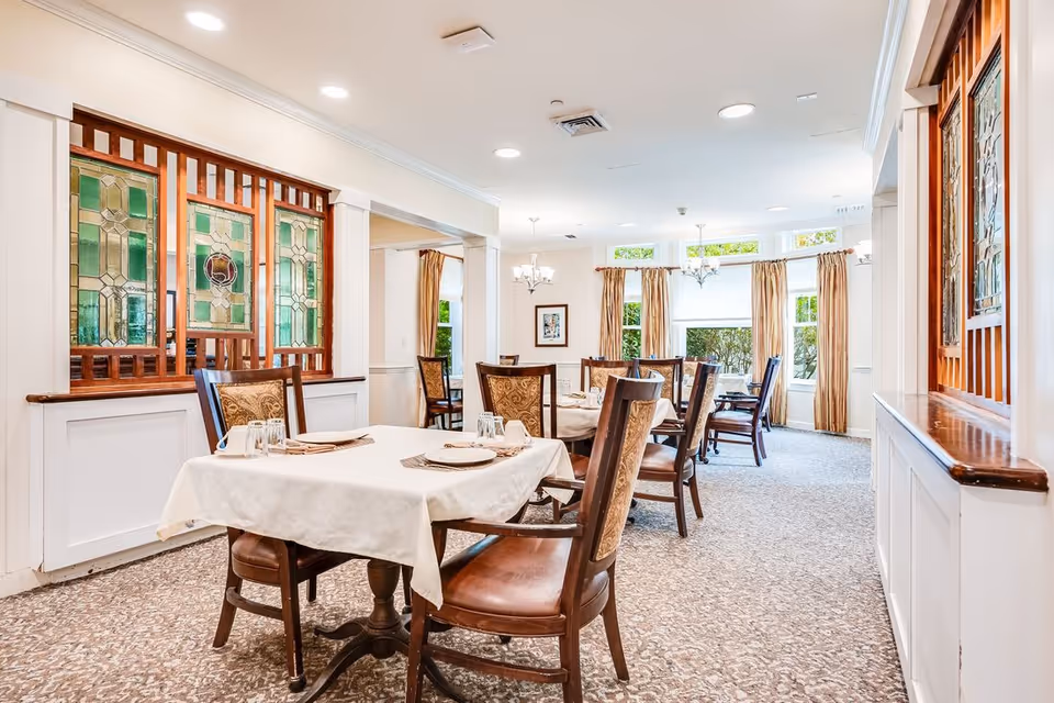 A bright dining room with several tables covered with white tablecloths, set with plates, glasses, and napkins. The room features wooden chairs with cushioned seats and backs, patterned carpet, large windows with beige curtains, and decorative stained glass panels on the walls.