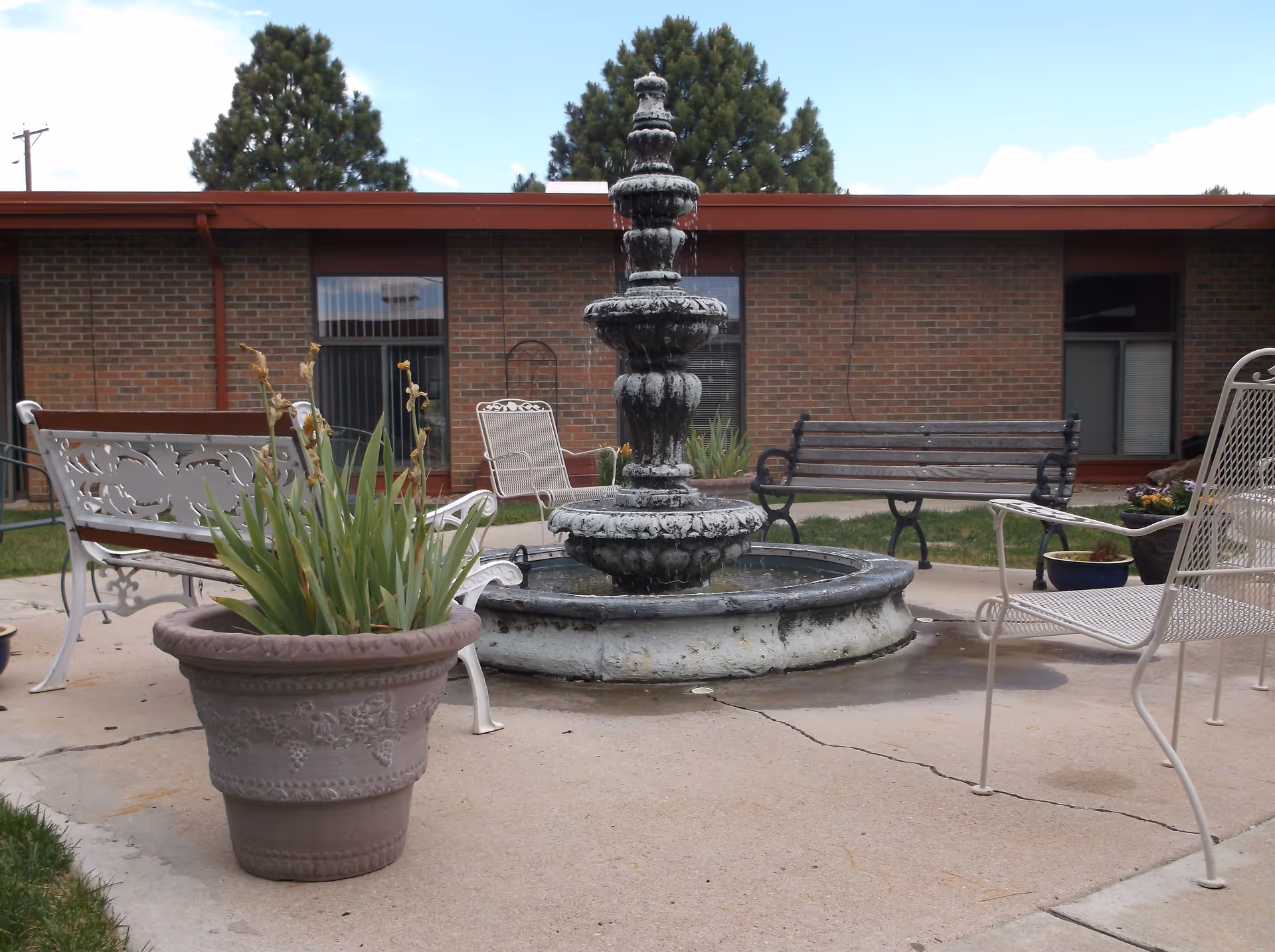 Courtyard with a tiered stone fountain surrounded by benches, metal chairs, and a potted plant in front of a brick building.
