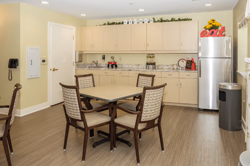 A small kitchen and dining area with a square table and four chairs in the center. The kitchen has beige cabinets, a marble-patterned countertop, a stainless steel refrigerator with a red 'eat' decoration on top, and a trash can nearby. The walls are light yellow, and there is a door and a wall-mounted telephone on the left side.