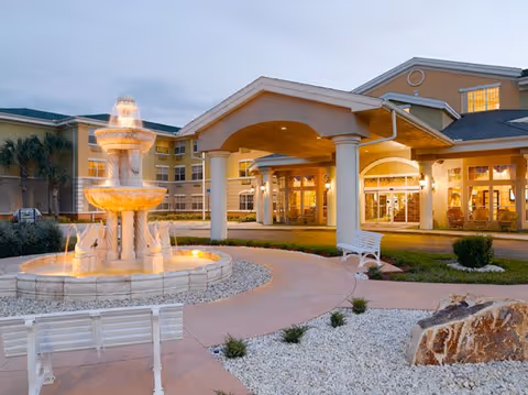 Entrance of a senior living building at dusk with a lit multi-tiered fountain, benches, and a covered porte-cochère.
