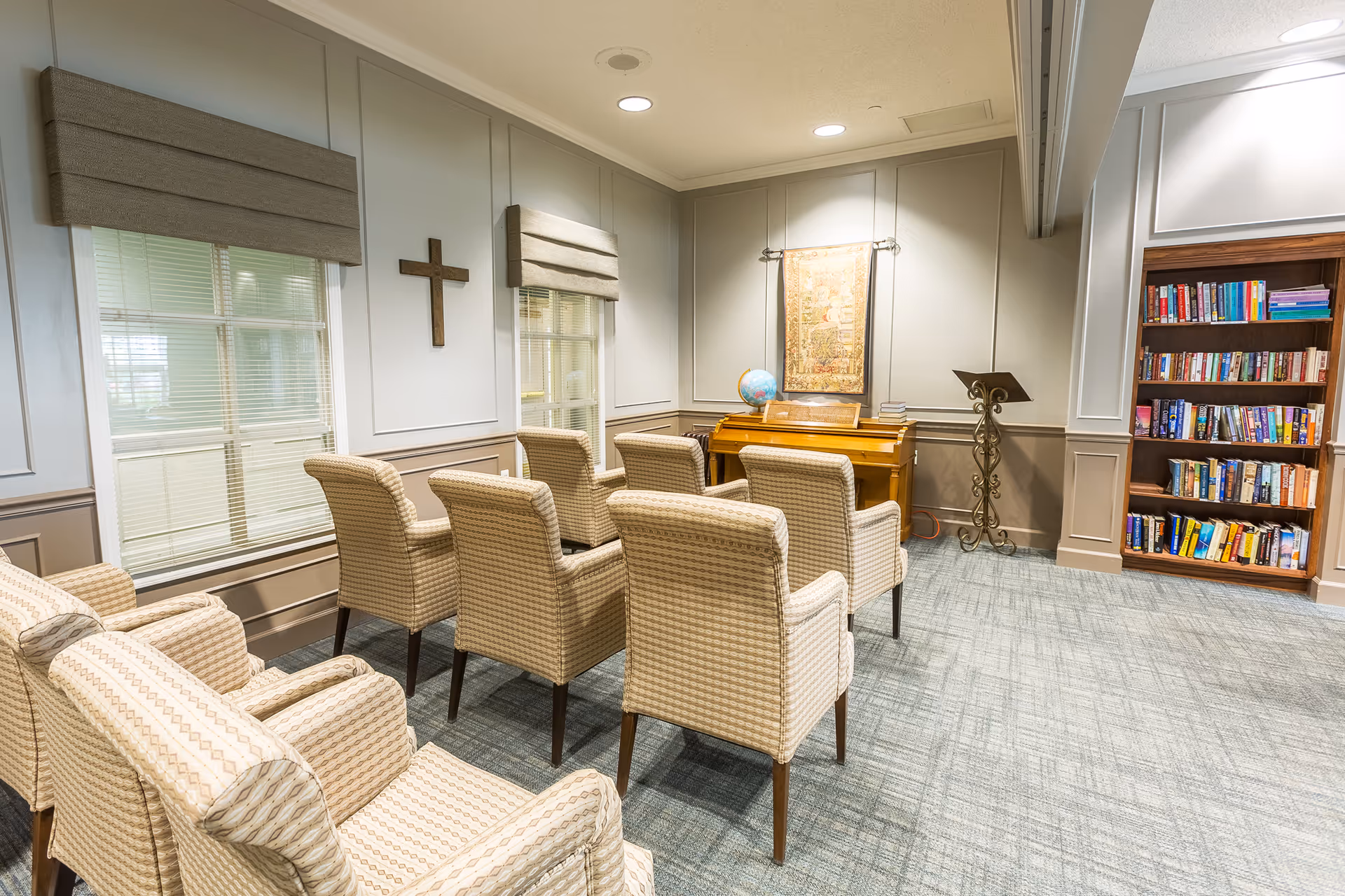 Cozy interior room with rows of upholstered chairs facing a piano and lectern, a bookshelf, and a wall cross.