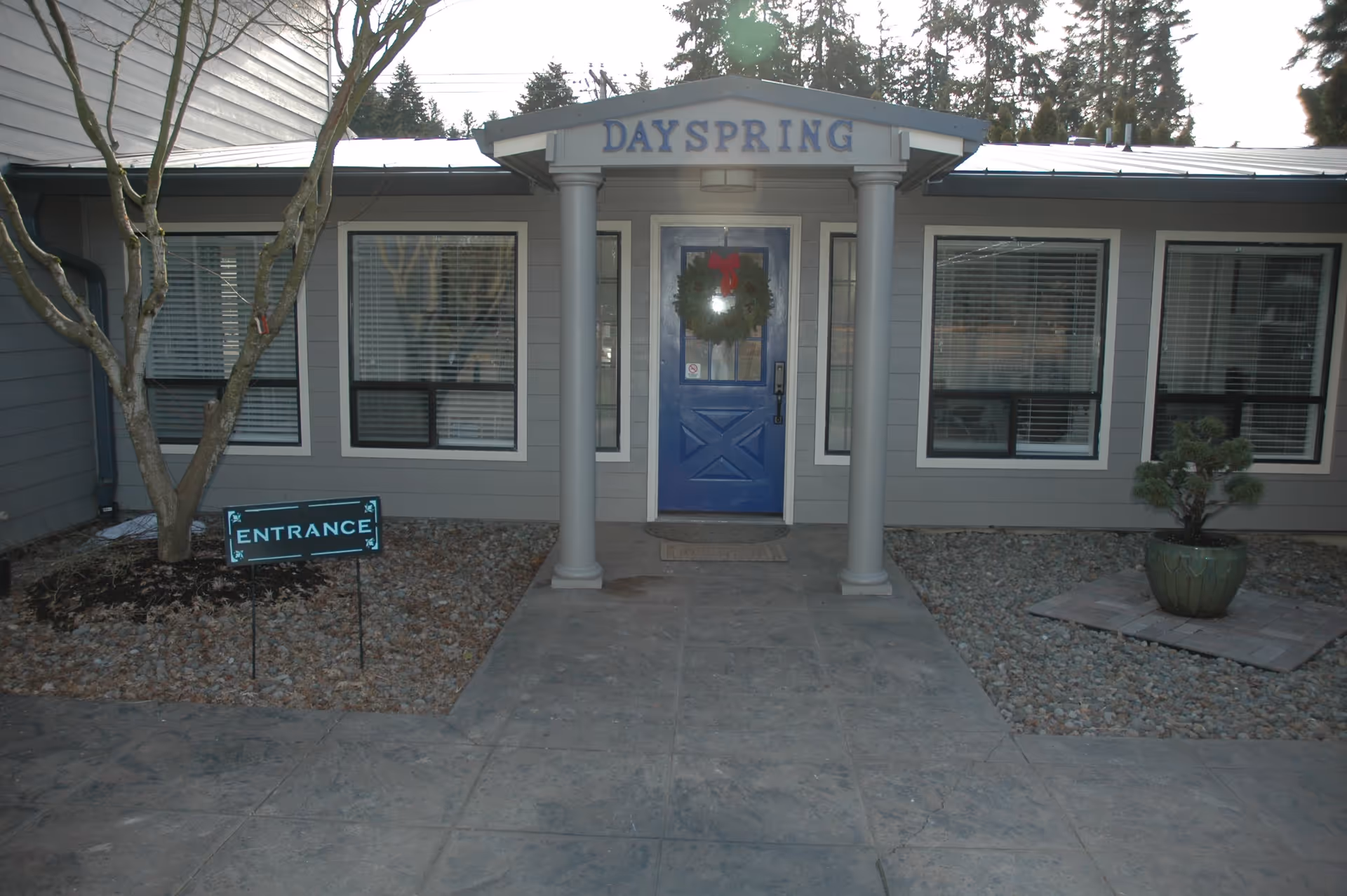 Front entrance of Dayspring Memory Care facility with a blue door decorated with a wreath, flanked by two columns and windows on either side. There is a sign in the garden bed to the left that says 'ENTRANCE'.